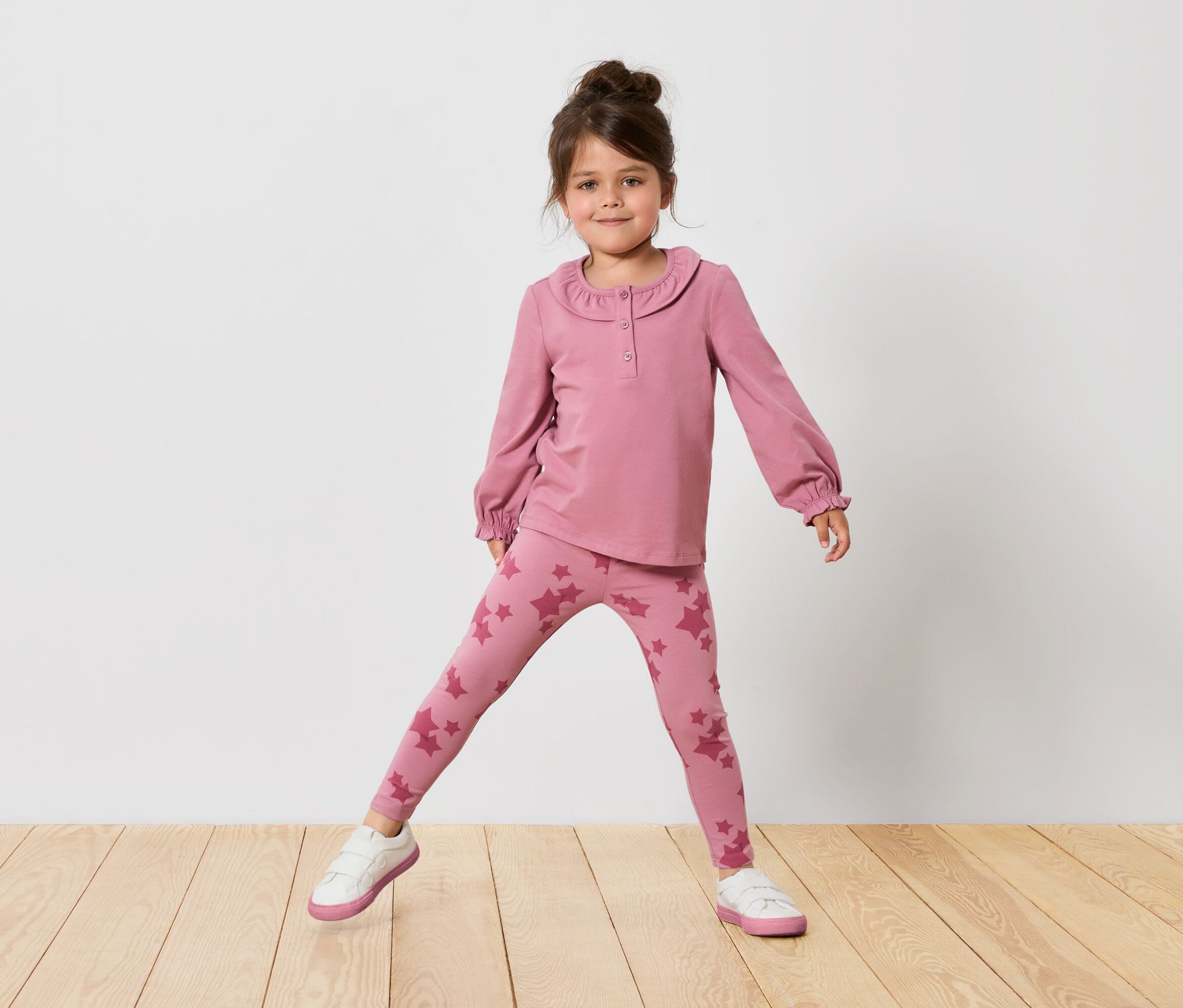 Dievča v ružovom tope s dlhým rukávom a legínach s hviezdami stojí na drevenej podlahe pred bielou stenou.
