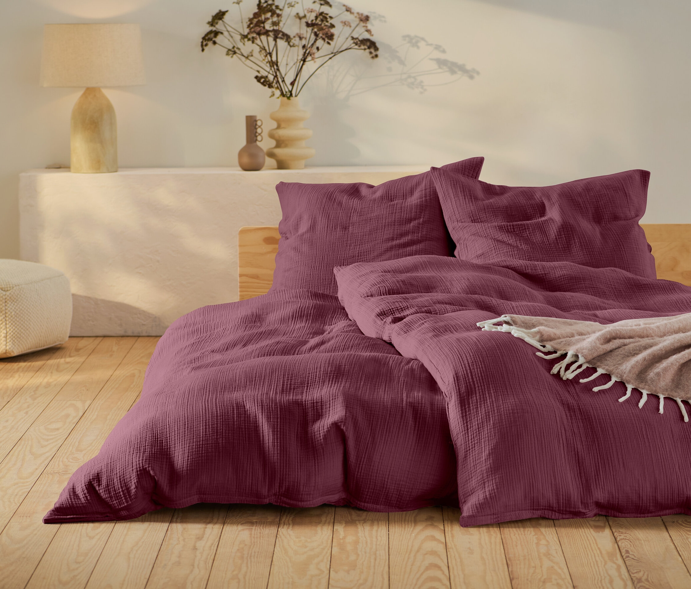 Vínová posteľná bielizeň na posteli s dreveným rámom, béžový puf a lampa na bielom stolíku.