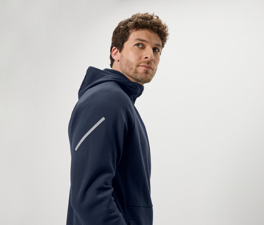 Muž s hnedými kučeravými vlasmi sa pozerá cez plece. Má na sebe tmavomodrú mikinu s kapucňou a diagonálny reflexný pruh na ramene.