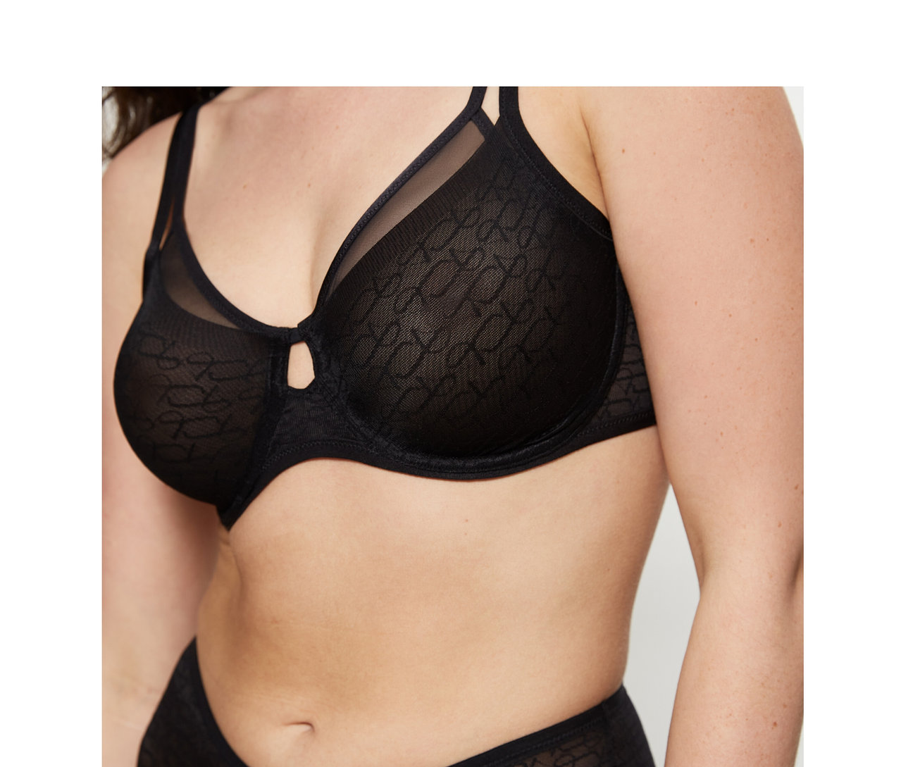Žena v čiernej podprsenke Triumph Signature Sheer Minimizer a zladených, vzorovaných nohavičkách. Podprsenka má malý výrez medzi košíčkami.