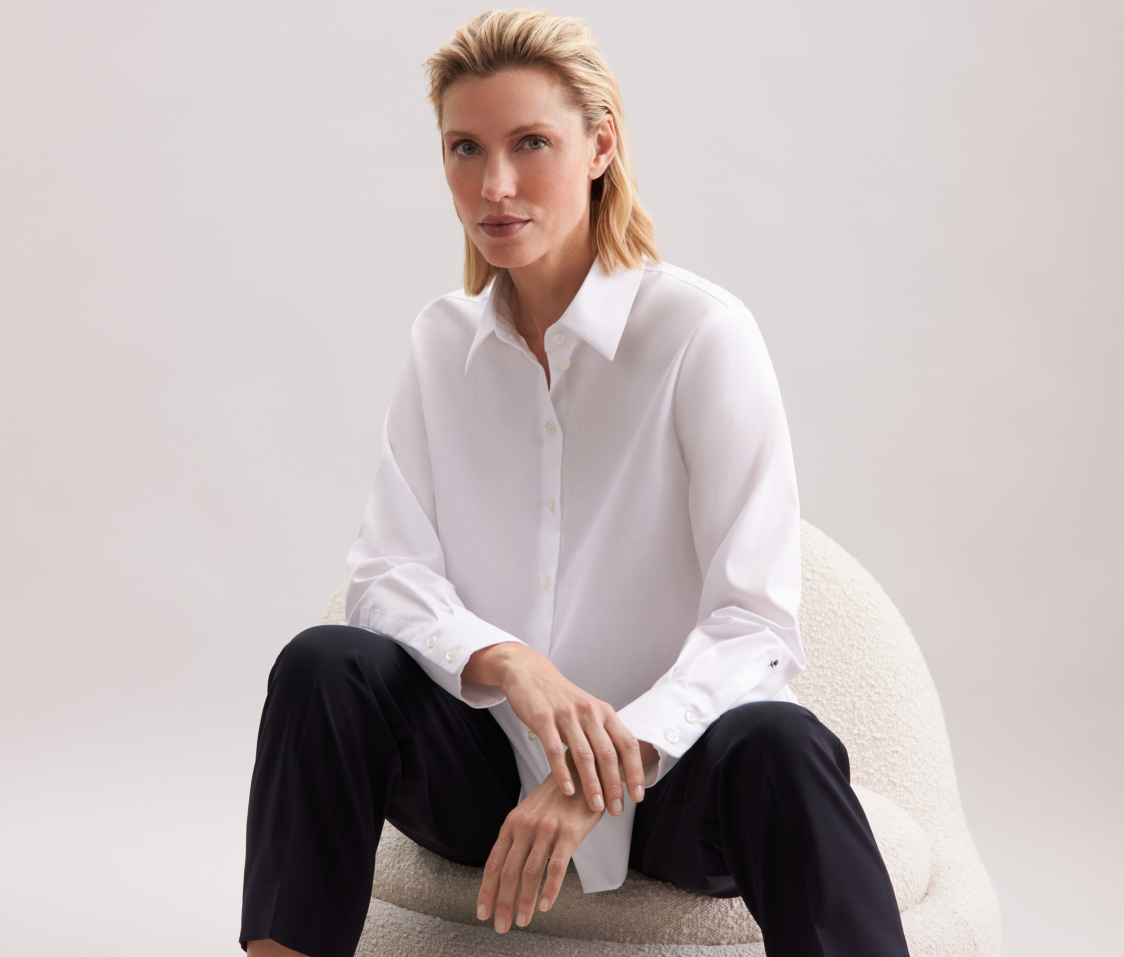 Žena sedí na stoličke a má na sebe bielu košeľu a čierne nohavice.