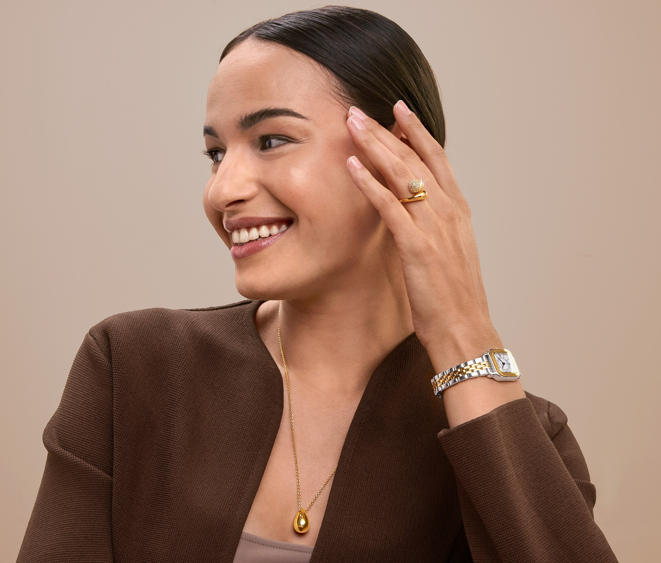 Usmievajúca sa žena sa dotýka tváre, má na sebe zlatý prsteň, hodinky a náhrdelník.