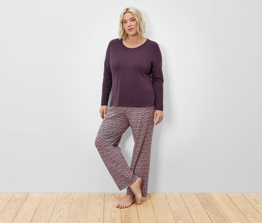 Blondínka v bordovom pyžame s dlhým rukávom a kvetinových nohaviciach pózuje pri stene na drevenej podlahe.