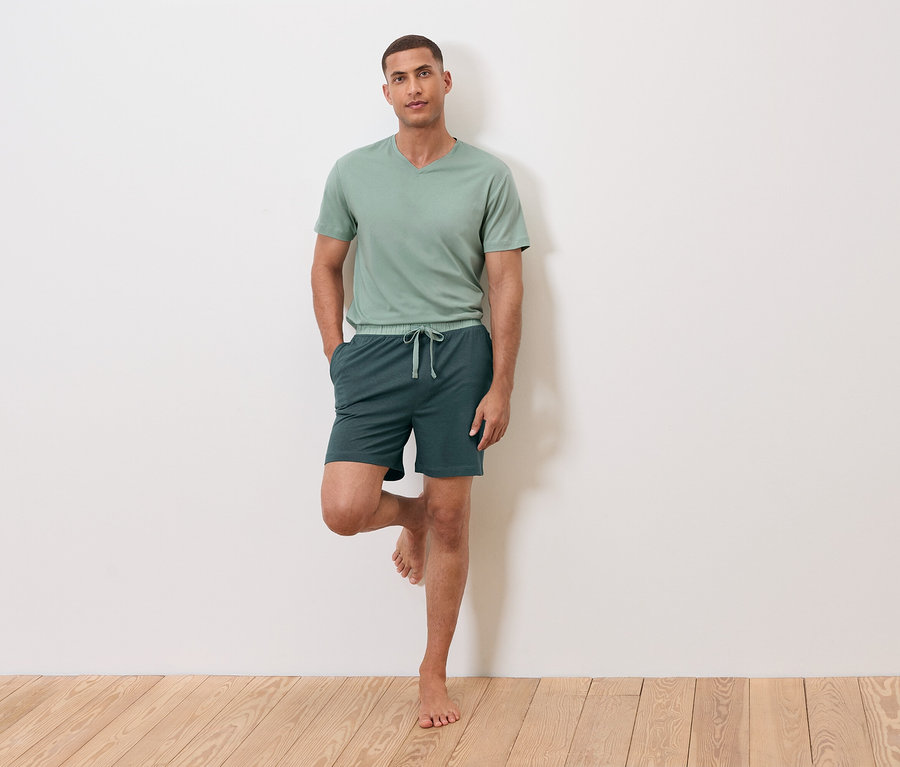 Muž stojí na jednej nohe opretý o stenu, má na sebe zelené tričko a šortky.