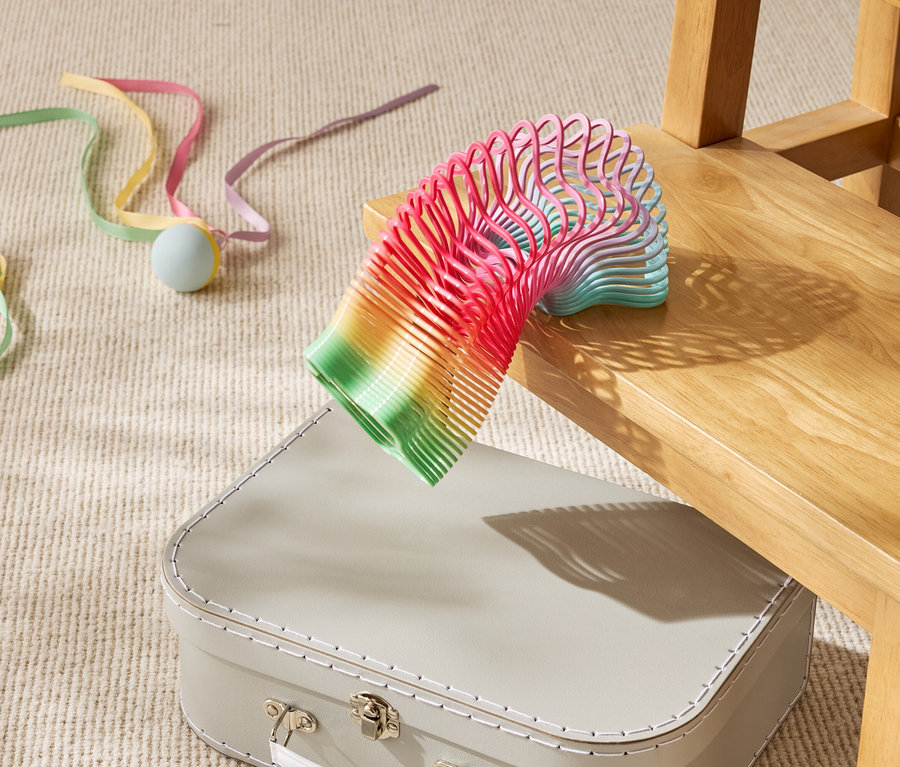 Farebná hračka špirála leží čiastočne na béžovom kufri pod drevenou stoličkou.