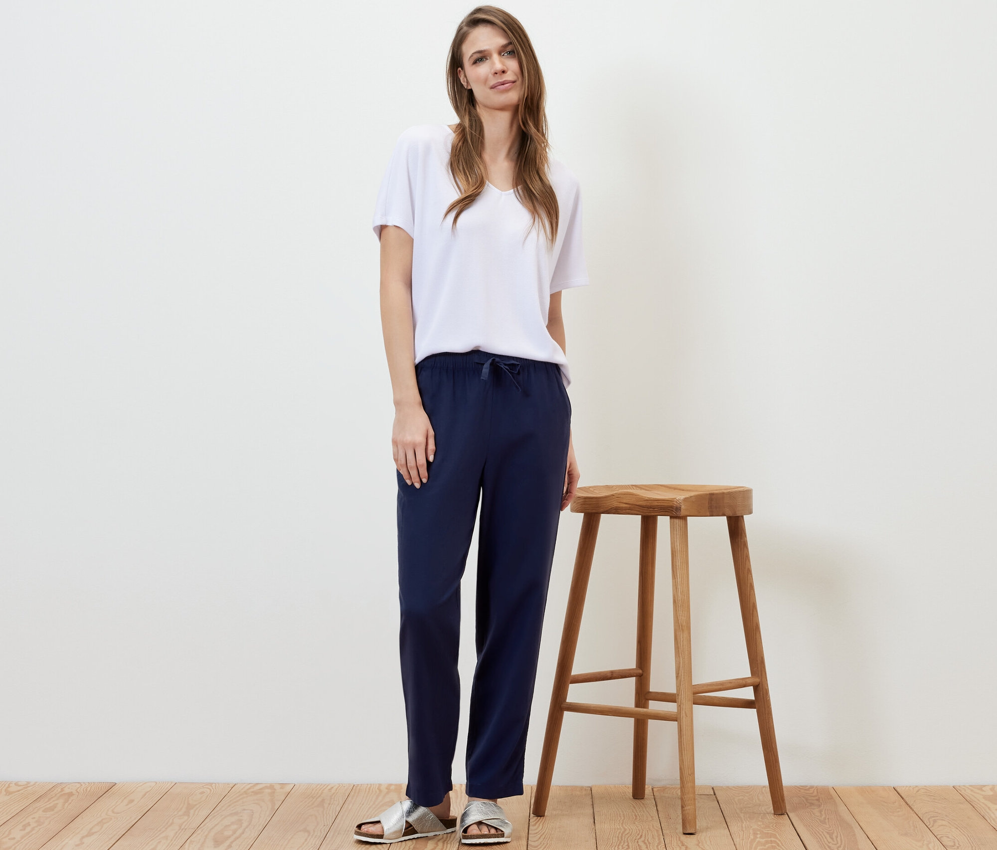 Žena stojí vedľa drevenej stoličky a má na sebe ľanové nohavice tmavomodrej farby a biele tričko.