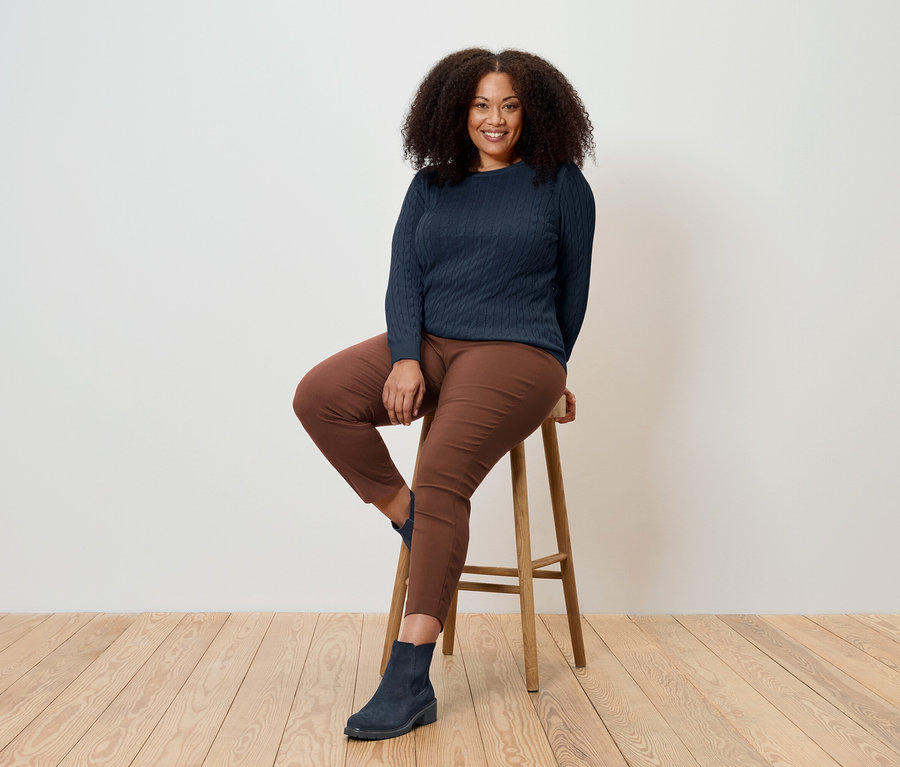 Žena sedí na drevenej stoličke, má na sebe tmavomodrý sveter, hnedé nohavice a modré topánky.