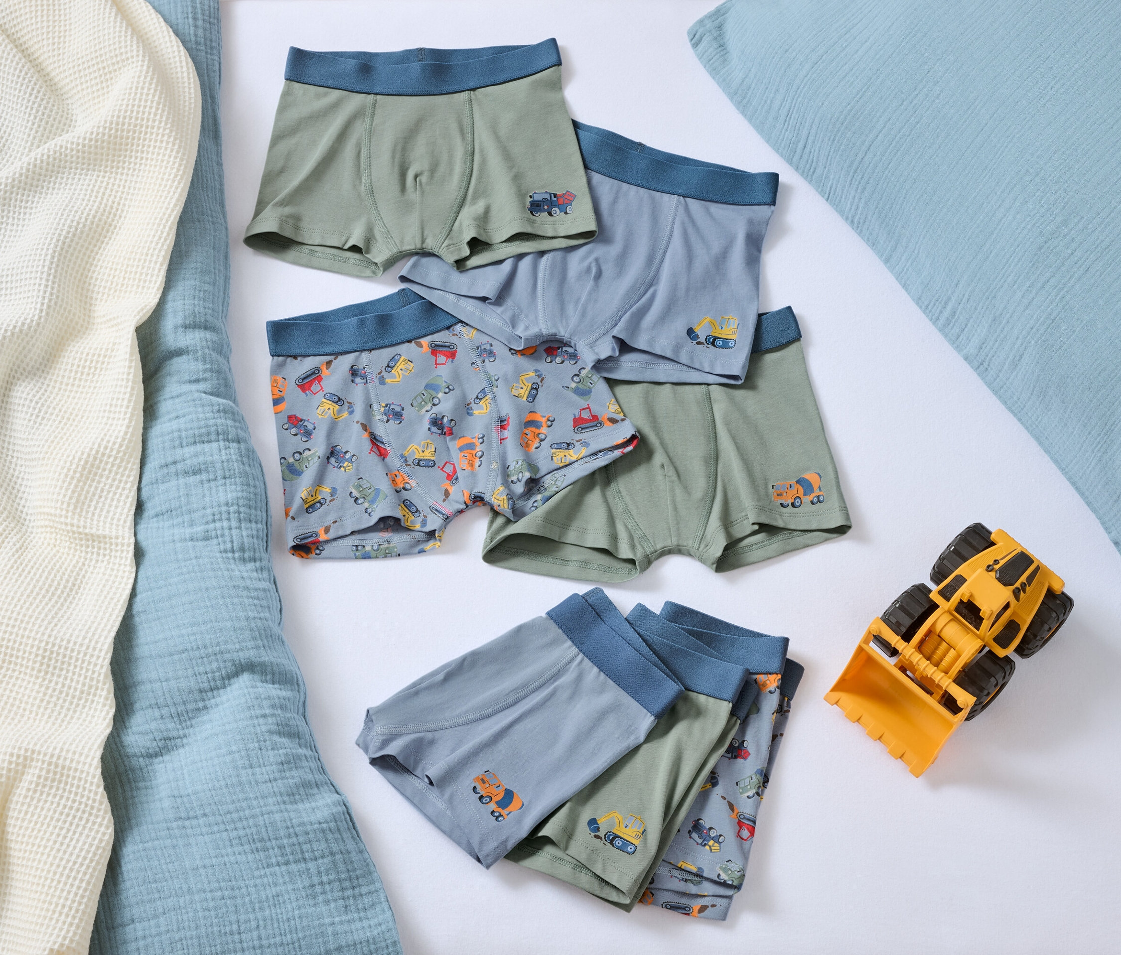 Na posteli leží hromada chlapčenských boxeriek s motívmi autíčok a žltá hračka buldozéra.