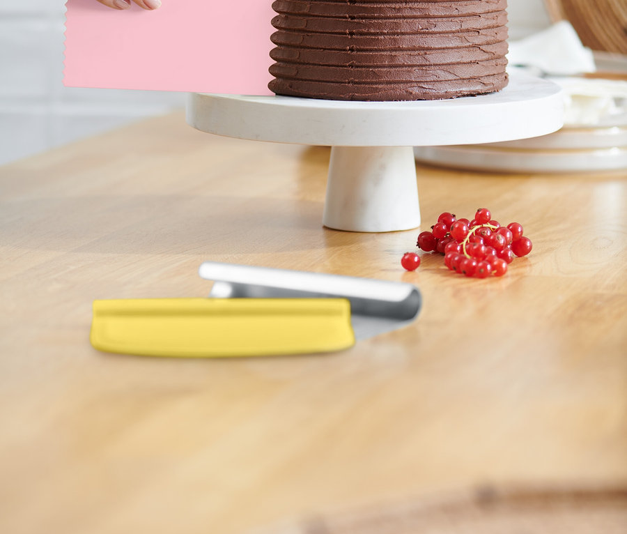 Ruka vyhladzuje polevu na čokoládovej torte ružovou špachtľou. Torta stojí na bielom podstavci a je ozdobená červenými ríbezľami. Na stole leží žltá špachtľa.