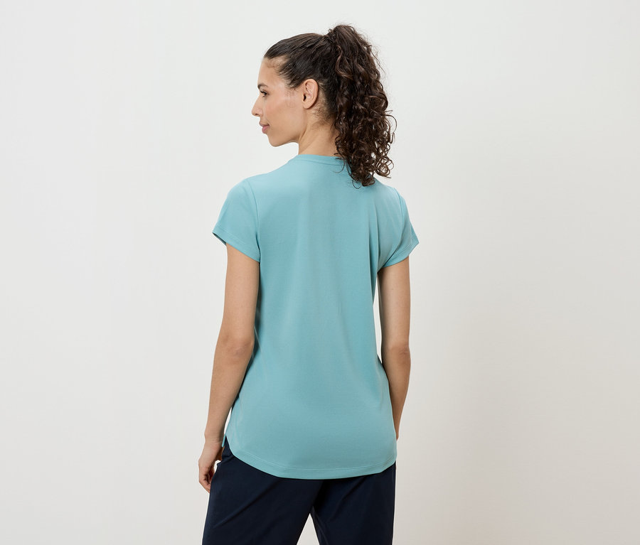 Žena s tmavými kučeravými vlasmi má na sebe tyrkysové tričko a tmavé nohavice, pohľad zozadu.