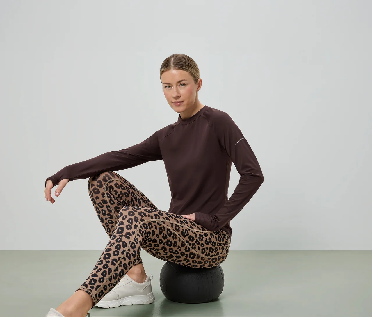 Žena sedí na čiernej lopte, má na sebe hnedú blúzku s dlhým rukávom, legíny s leopardím vzorom a biele tenisky.