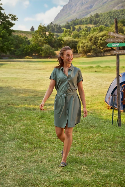 Žena kráča po tráve v zelených košeľových šatách s opaskom. V pozadí je vidieť stan a smerovník.
