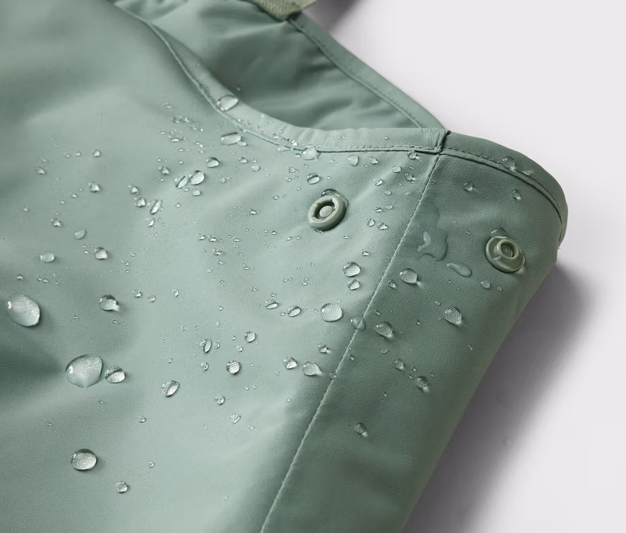 Detailný záber mätovo zeleného, vodeodolného materiálu s patentkami a kvapkami vody.