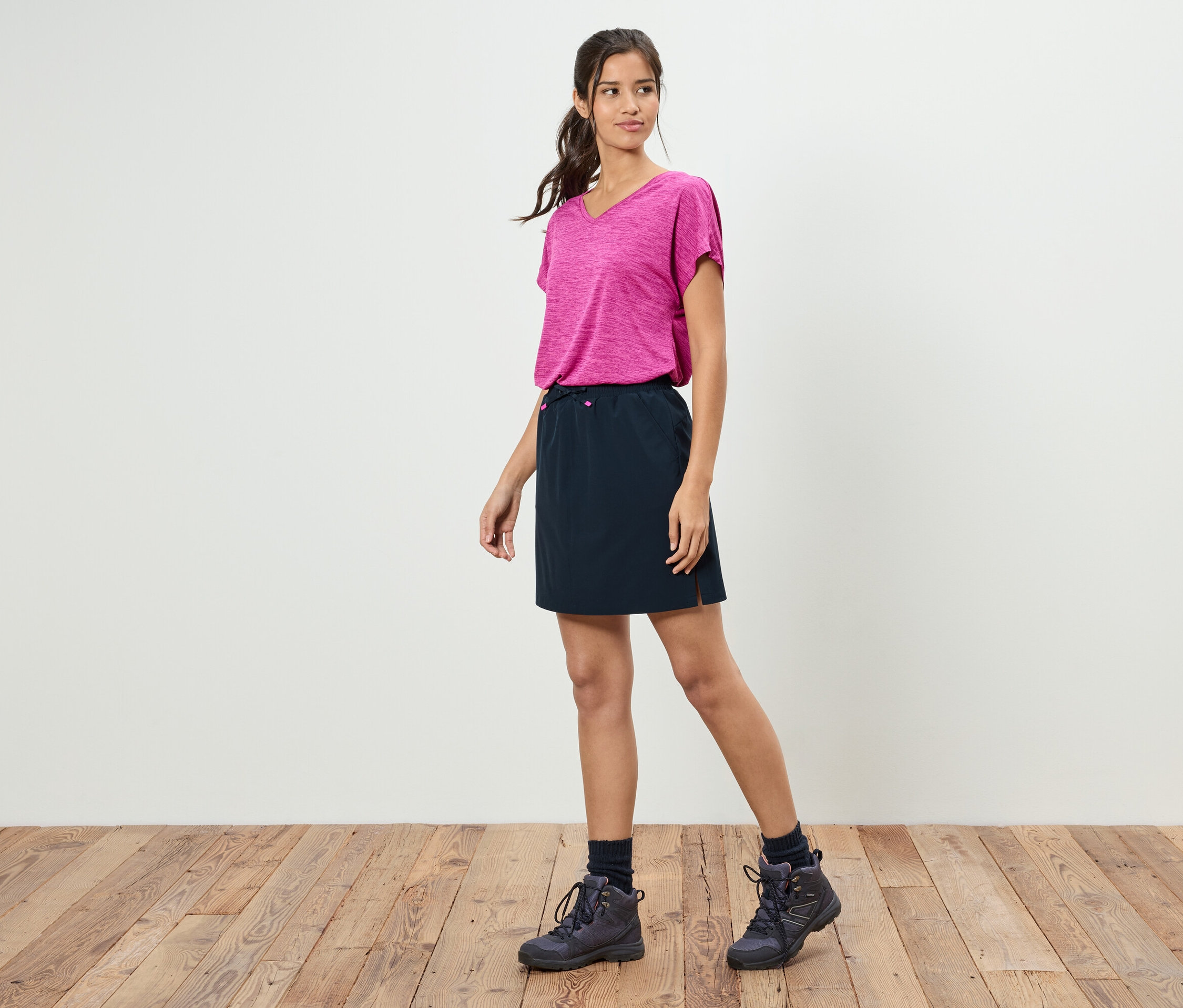 Žena stojí pred bielou stenou a má na sebe ružové tričko, čiernu sukňu a turistické topánky.