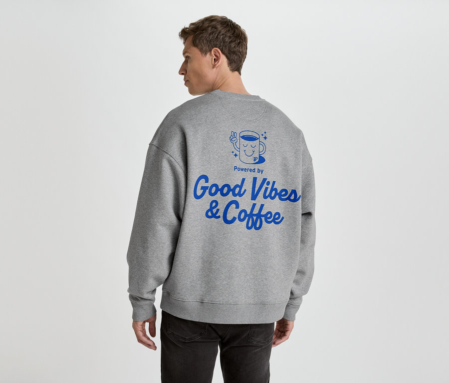 Muž zozadu v šedej mikine s modrou potlačou 'Good Vibes & Coffee'.