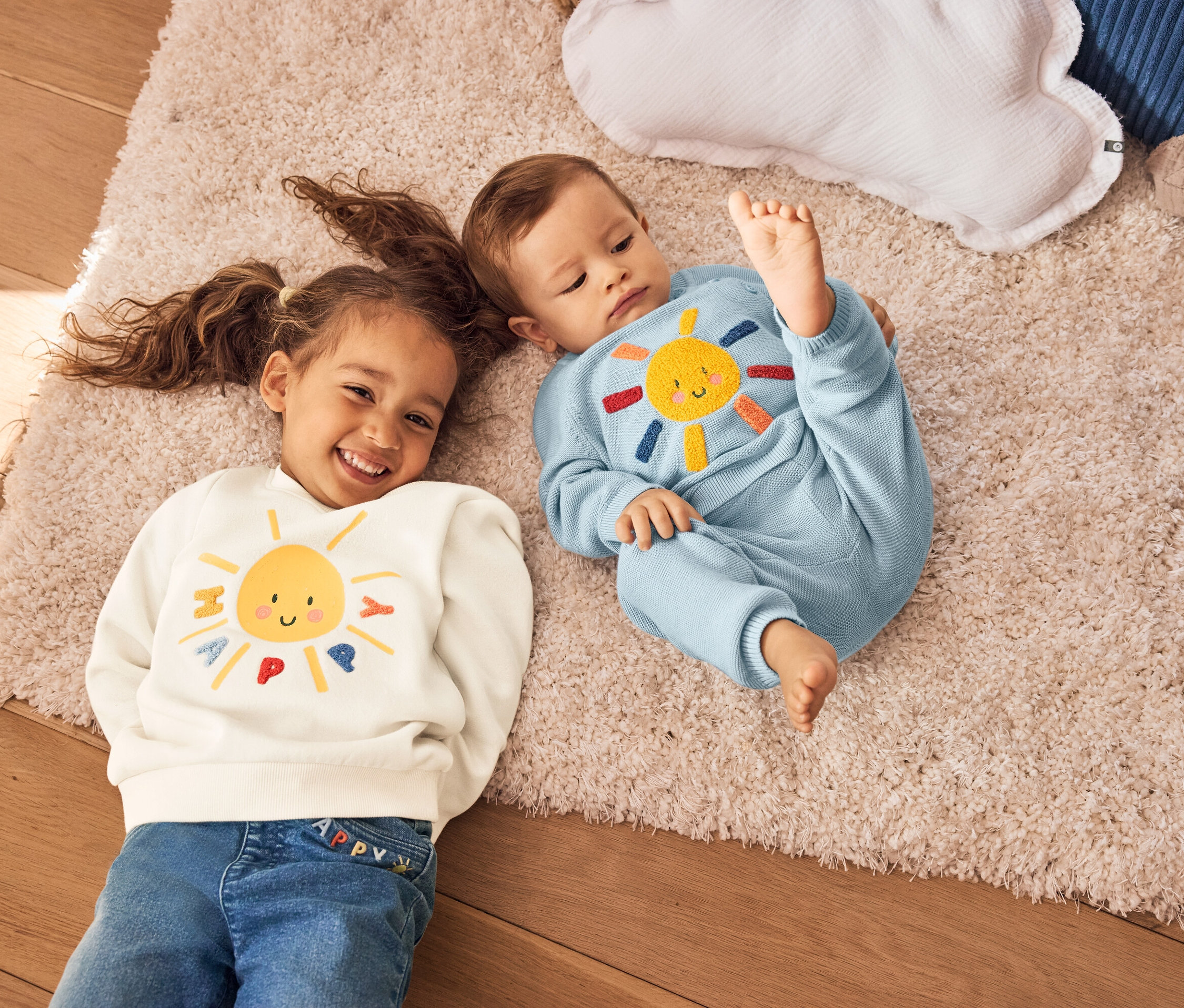 Dievča s vrkočmi a dieťa ležia na koberci. Dievča má na sebe krémovú mikinu so žltým slnkom. Dieťa má na sebe modrý pletený oblek so slnkom.