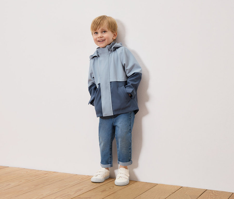 Usmievavý chlapec s blond vlasmi stojí pred bielou stenou a má na sebe dvojfarebnú, svetlomodrú a námornícku bundu, modré džínsy a biele tenisky.
