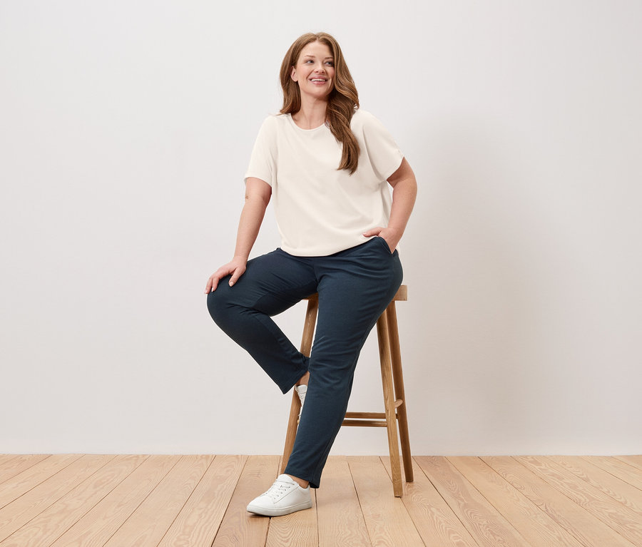 Žena sedí na stoličke, má na sebe krémové tričko, modré nohavice a biele tenisky.