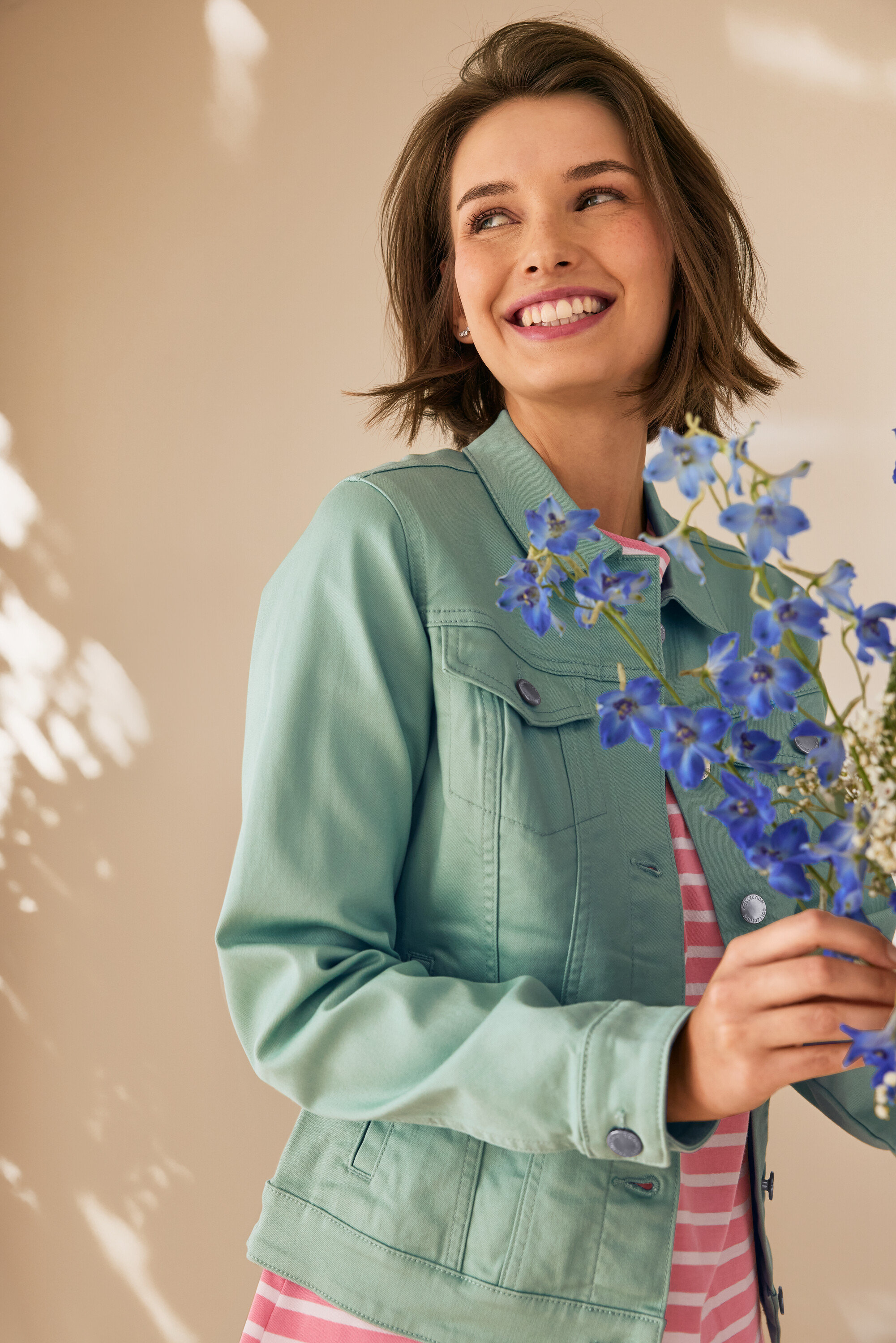 Žena sa usmieva a drží modré kvety, má na sebe mätovozelenú džínsovú bundu.