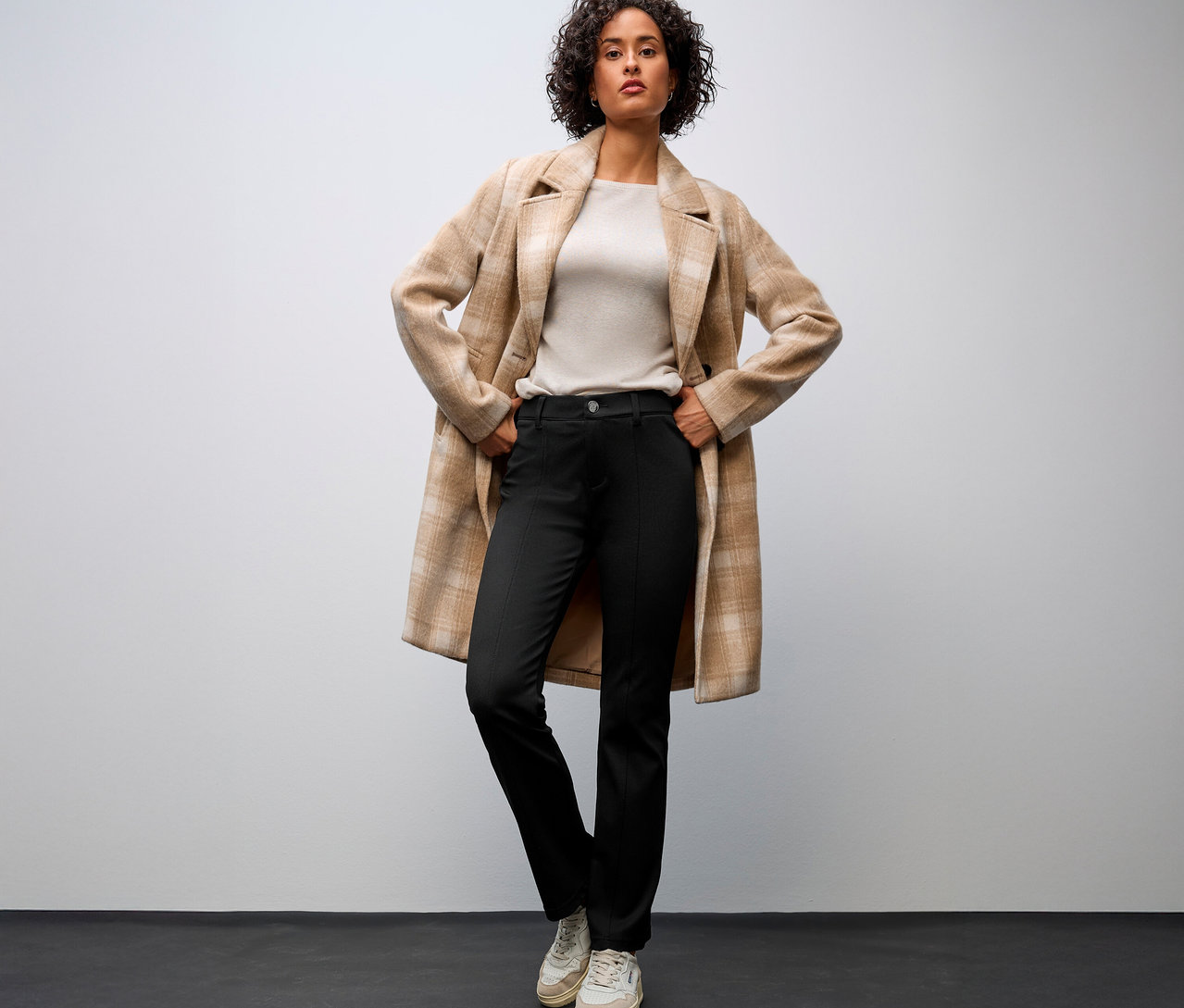 Žena v béžovom kabáte a Street One Bootcut nohaviciach s rukami na bokoch.