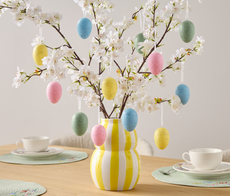 Dekoratívne kraslice visia na kvitnúcich konároch vo váze so žlto-bielymi pruhmi, stojacej na stole s prestieraním.