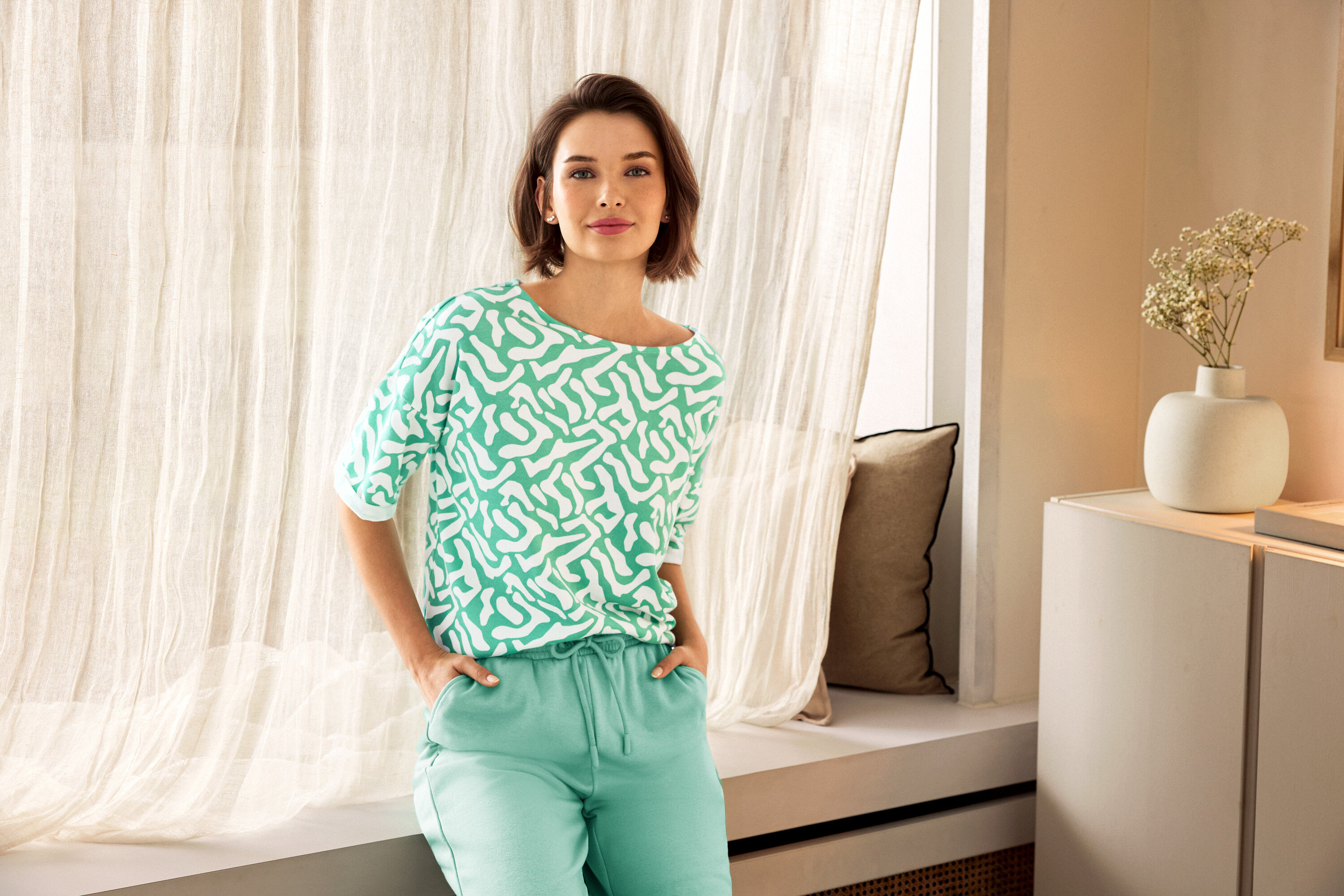 Žena v zelených nohaviciach a vzorovanej blúzke pózuje vedľa okna. V pozadí je vidieť váza.