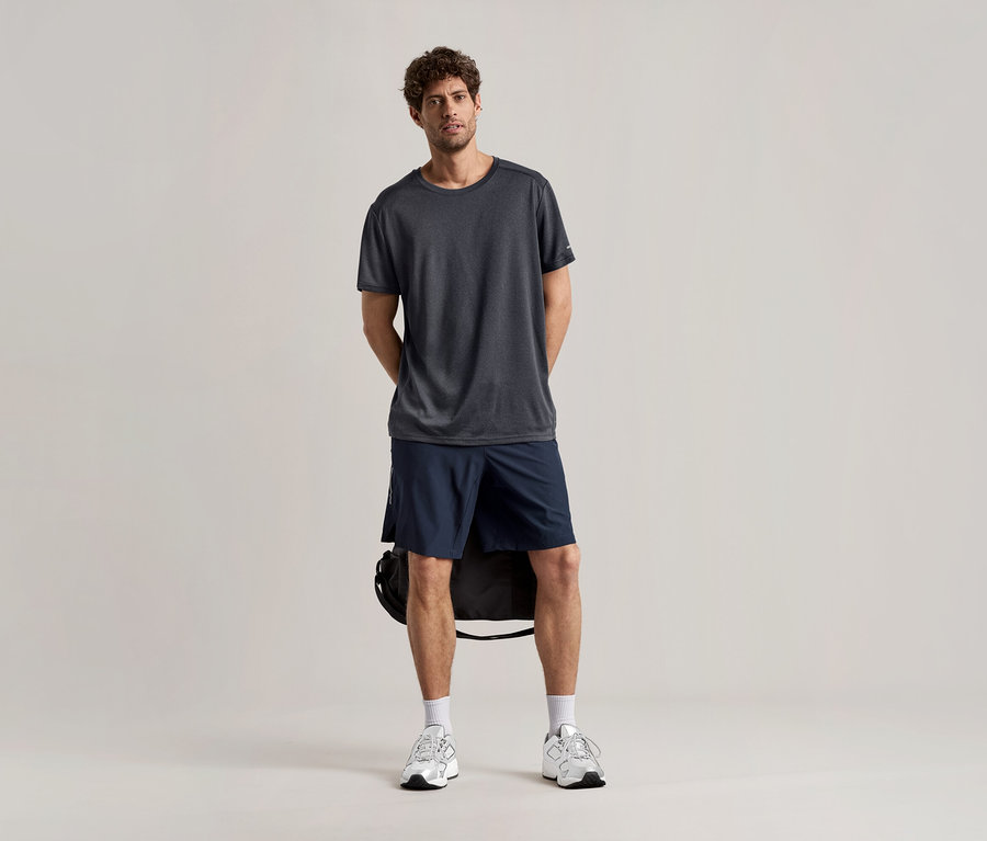 Muž stojí na svetlom pozadí v športovom tričku antracitovej farby, v šortkách a teniskách.