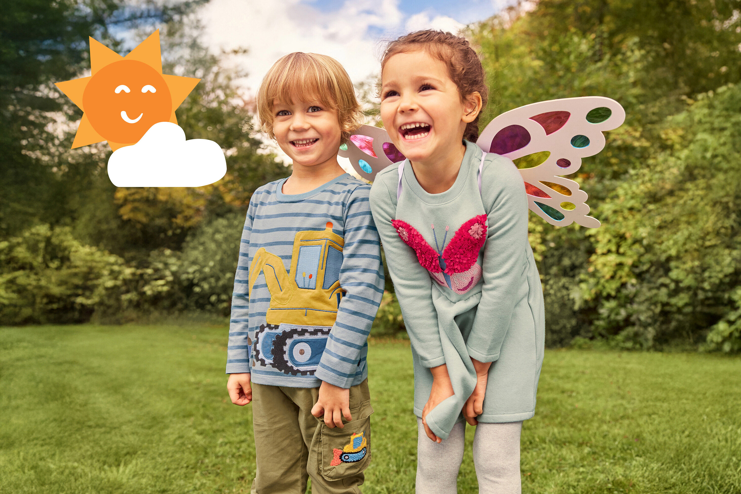 Dve smejúce sa deti stoja v tráve. Dievča má na sebe krídla a šaty s motýľom, chlapec má tričko s bagrom.