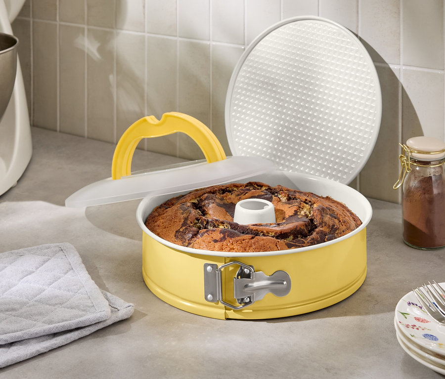 Mramorový koláč v žltej tortovej forme s vrchnákom a zdvíhacou rukoväťou vedľa kuchynského robota a pohára s korením