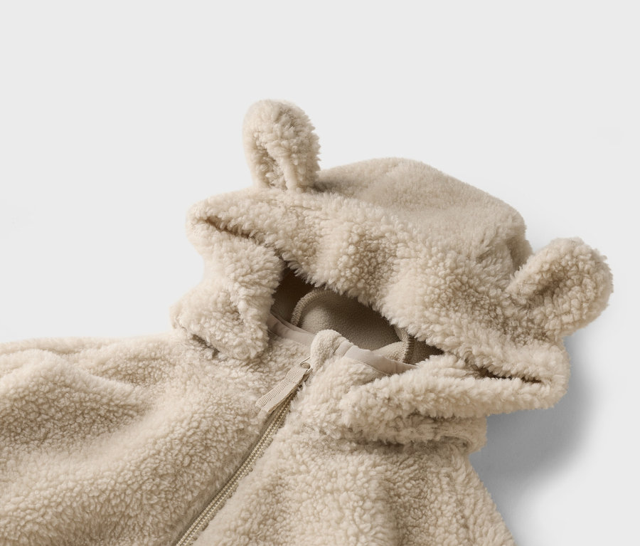 Detail béžovej, huňatej bundy s kapucňou a ušami na bielom pozadí.