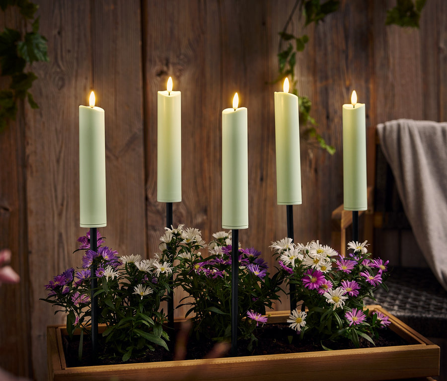 Päť horiacich, zelených sviečok stojí v drevenej krabici s fialovými a bielymi kvetmi pred drevenou stenou.