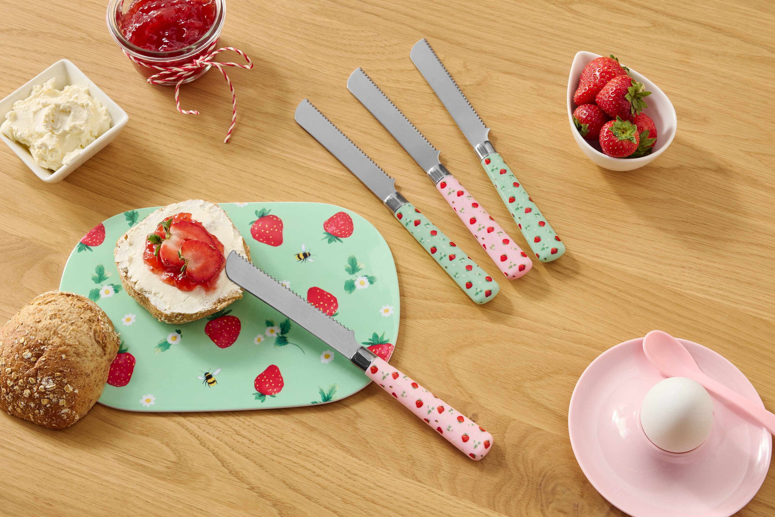 Na drevenom stole sa nachádza žemľa, tanier so vzorom jahôd, smotanový syr, džem, jahody a štyri nože so vzorom jahôd.