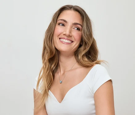 Žena s vlnitými hnedými vlasmi a bielym topom sa usmieva a má na sebe strieborný náhrdelník s modrým príveskom.