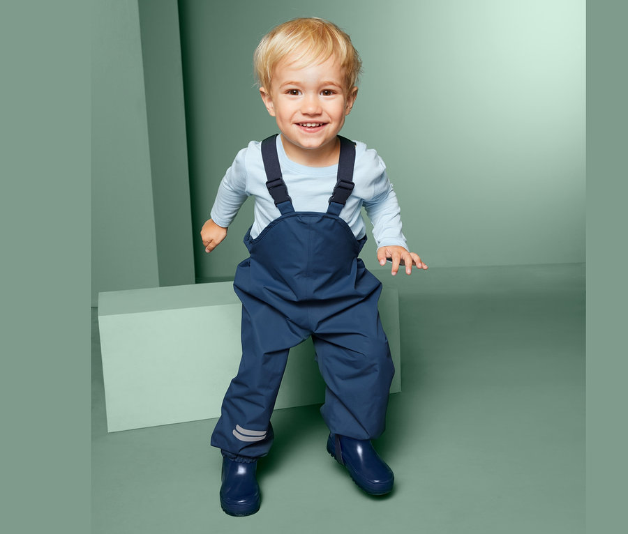 Usmievajúce sa batoľa stojí a má na sebe modré nohavice do dažďa s náprsenkou a modré gumáky.