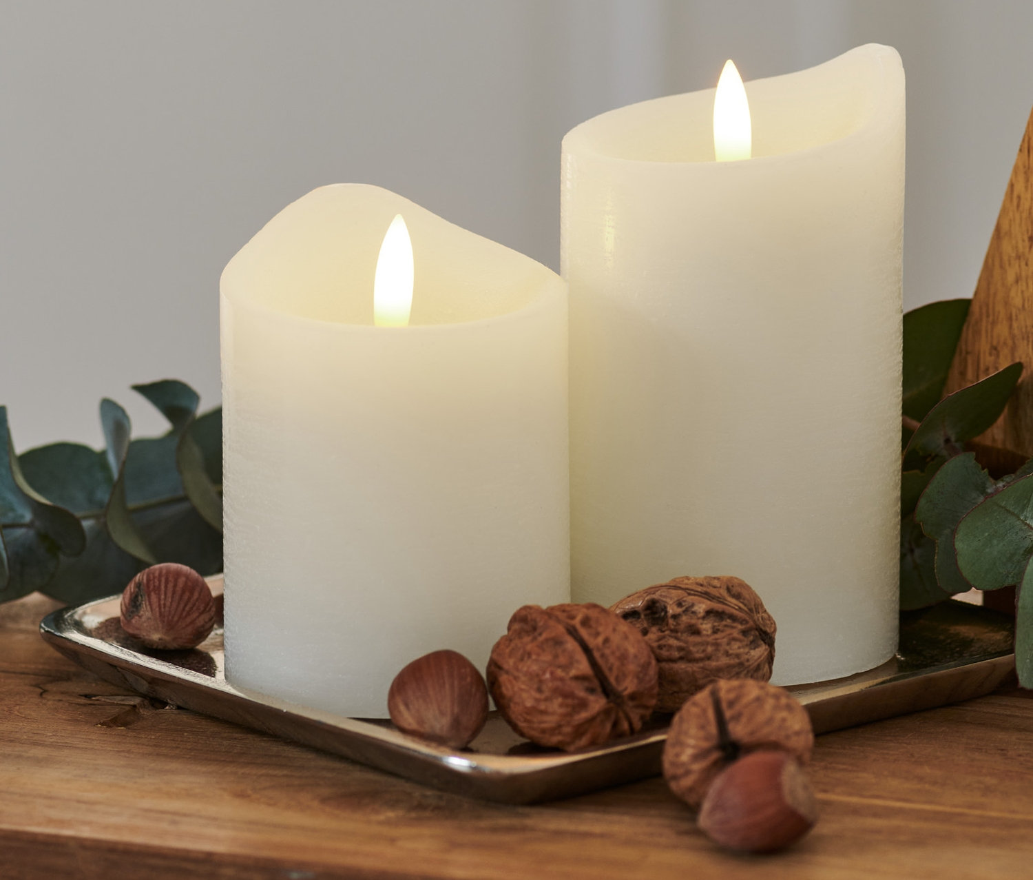 Na drevenom stole ležia na striebornej tácke dve biele LED sviečky z pravého vosku, orechy a vetvička eukalyptu.