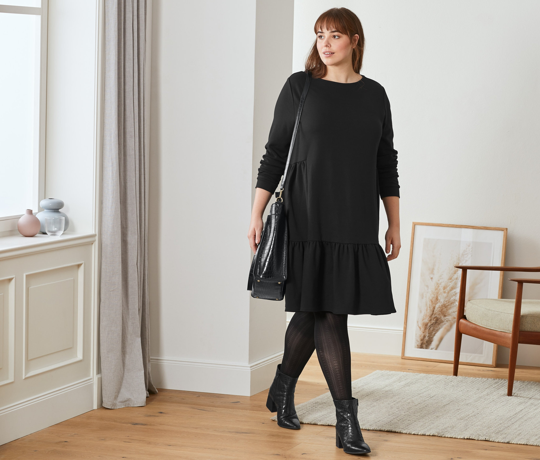 Žena kráča a má na sebe čierne teplákové šaty s volánom, členkové kožené čižmy so štruktúrou a pančuchové nohavice s ažúrovým vzorom. Cez rameno má prehodenú čiernu dámsku kabelku.