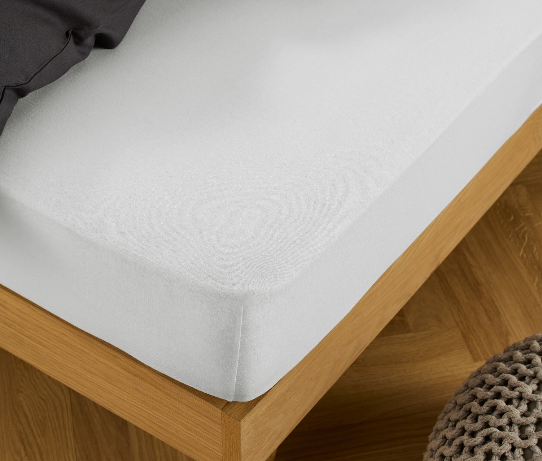 Biele džersejové napínacie prestieradlo je natiahnuté na posteli s dreveným rámom, vedľa stojí pletený puf.