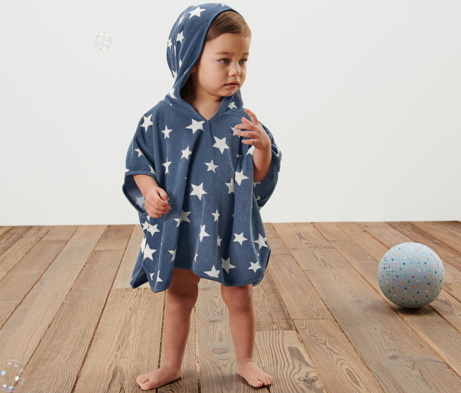 Dieťa stojí na drevenej podlahe a má na sebe modré froté pončo s kapucňou a bielymi hviezdami.