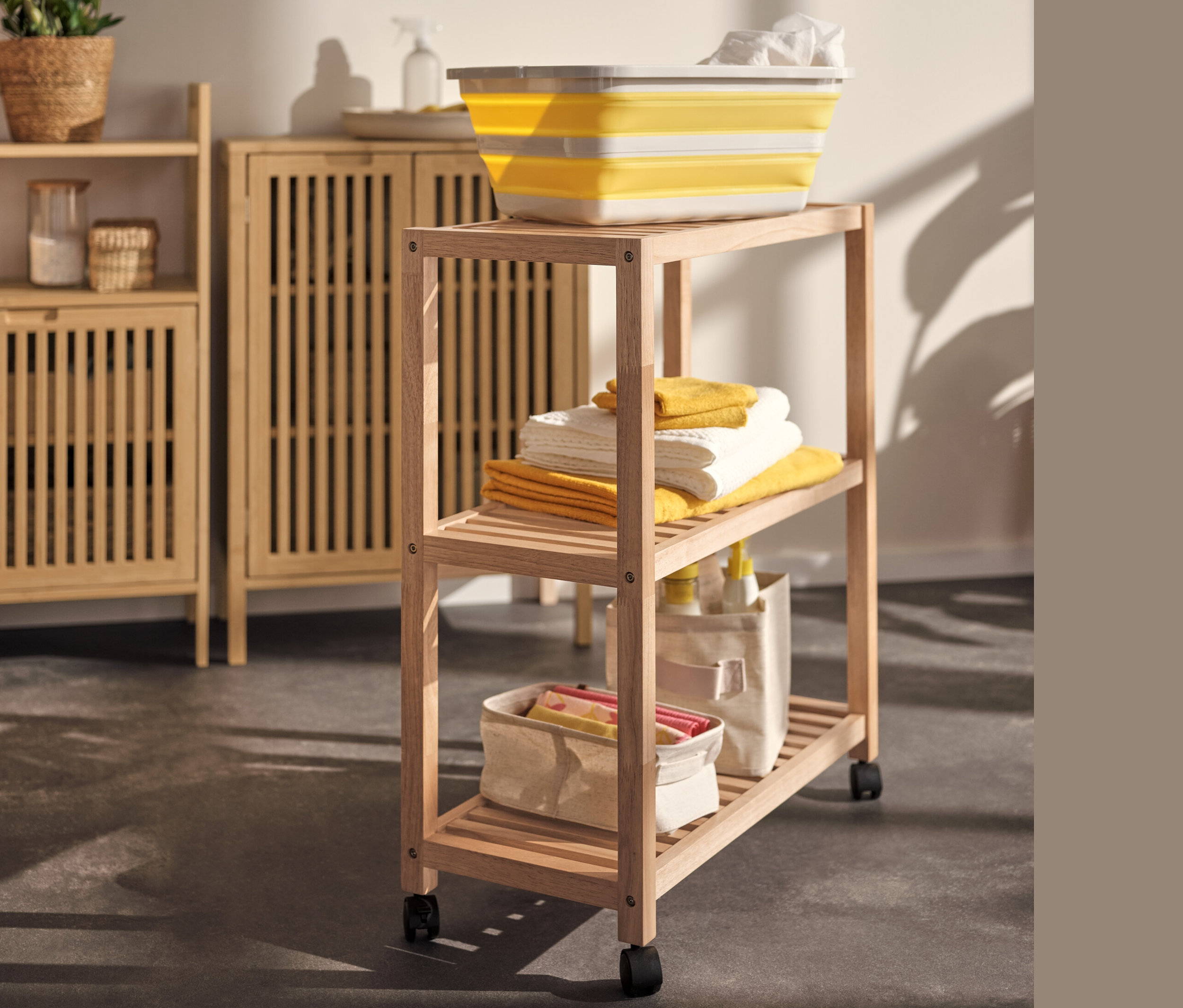 Žltý skladací kôš na bielizeň na regáli z masívneho dreva s kolieskami vedľa odkladacej skrinky.