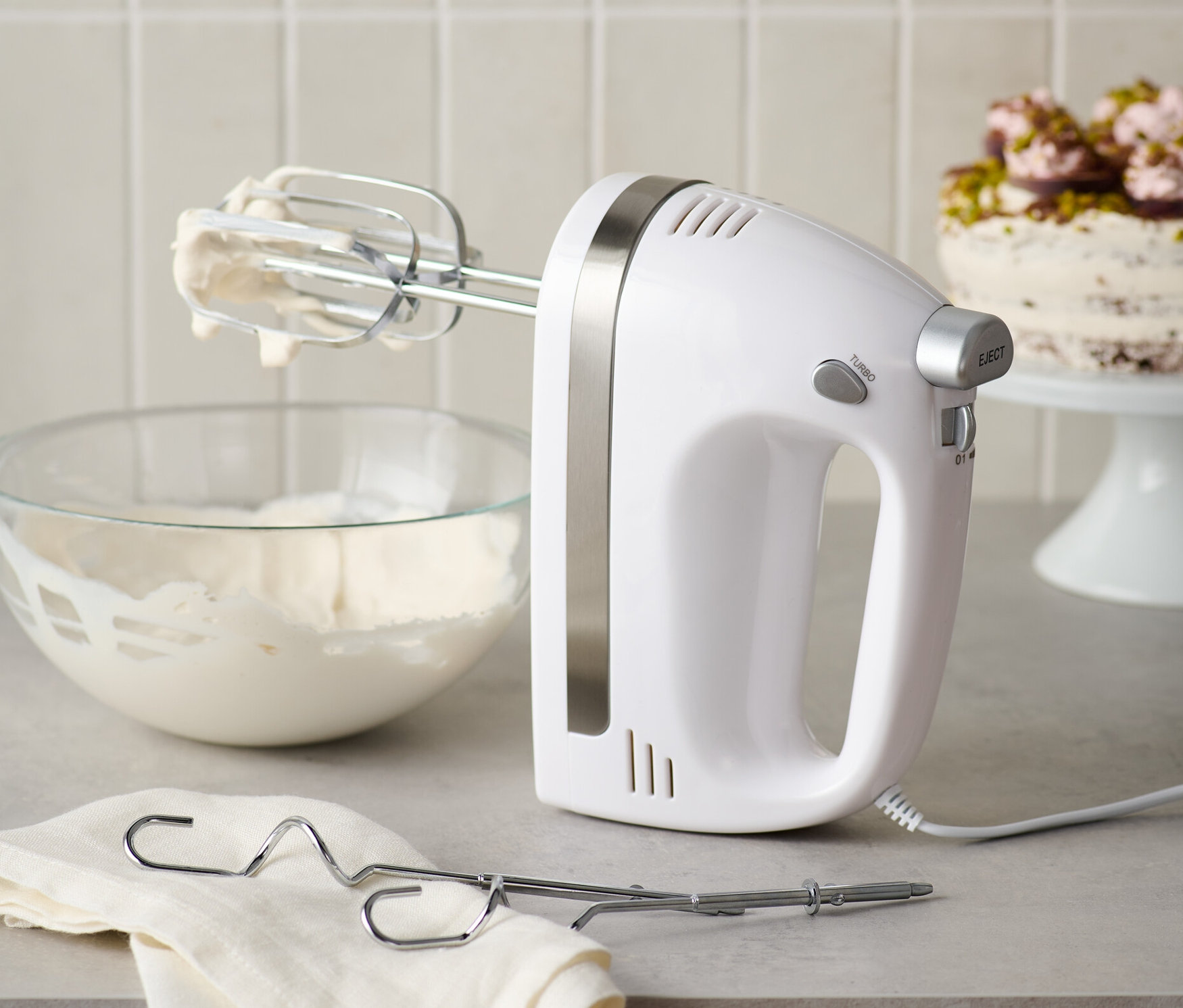 Biely ručný mixér s nerezovými metličkami stojí vedľa misy so šľahačkou a handričky.