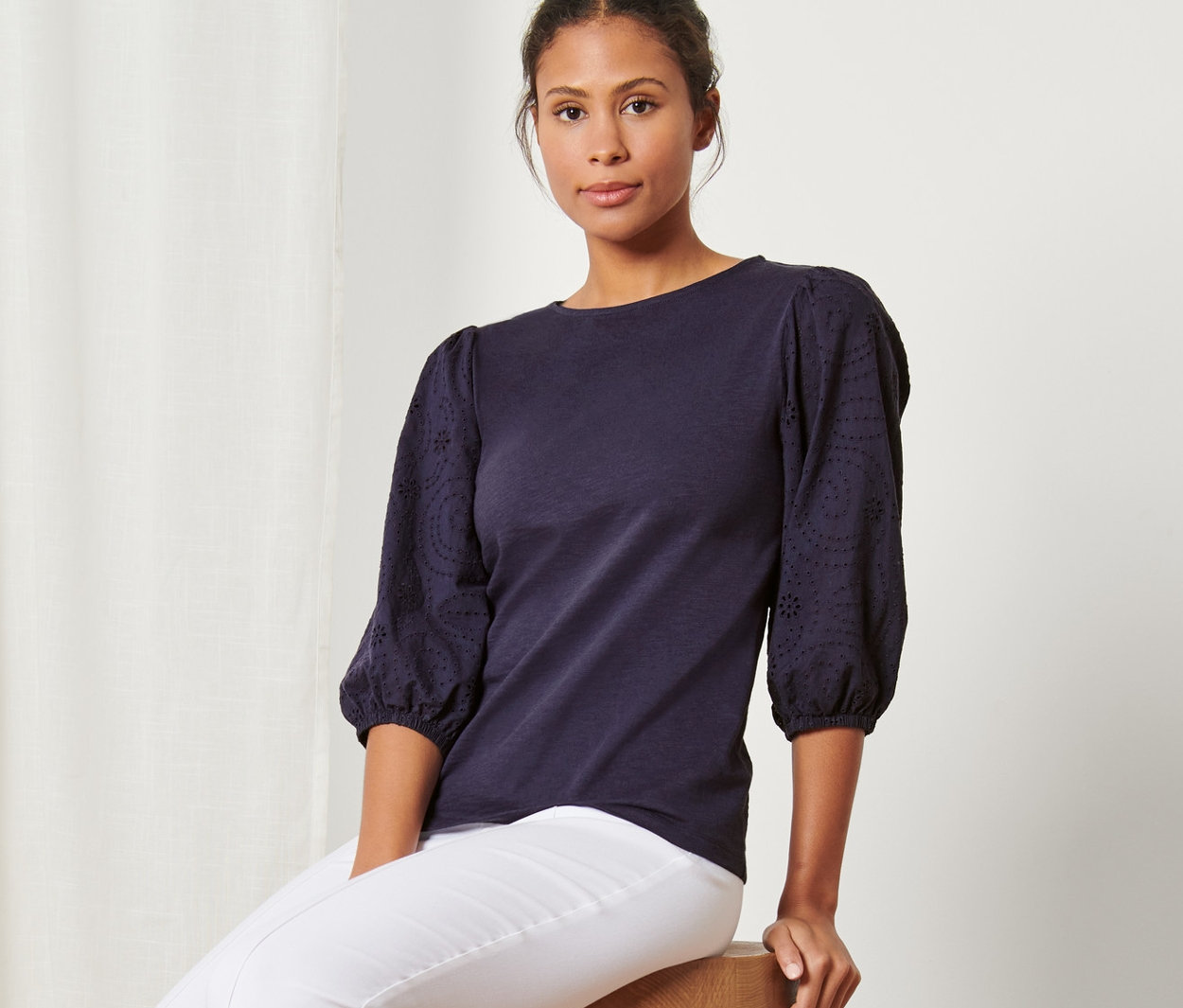 Žena sedí na stoličke a má na sebe tričko s ažúrovou výšivkou, biele nohavice a tenisky.