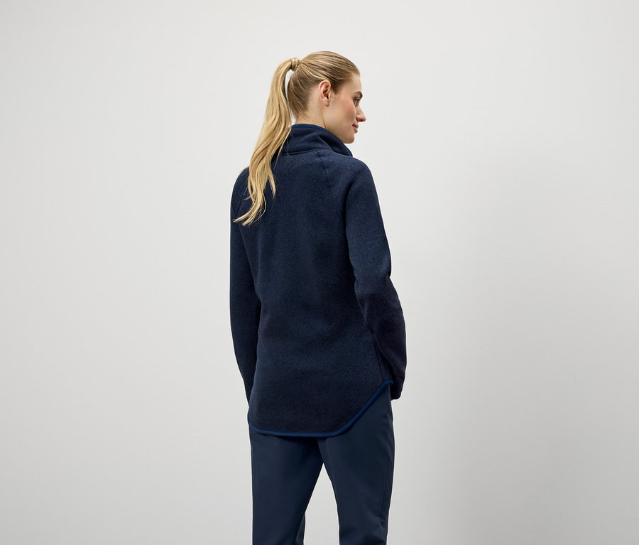 Zozadu je vidieť žena s blond vlasmi stiahnutými do copu, oblečená v tmavomodrom svetri a nohaviciach.