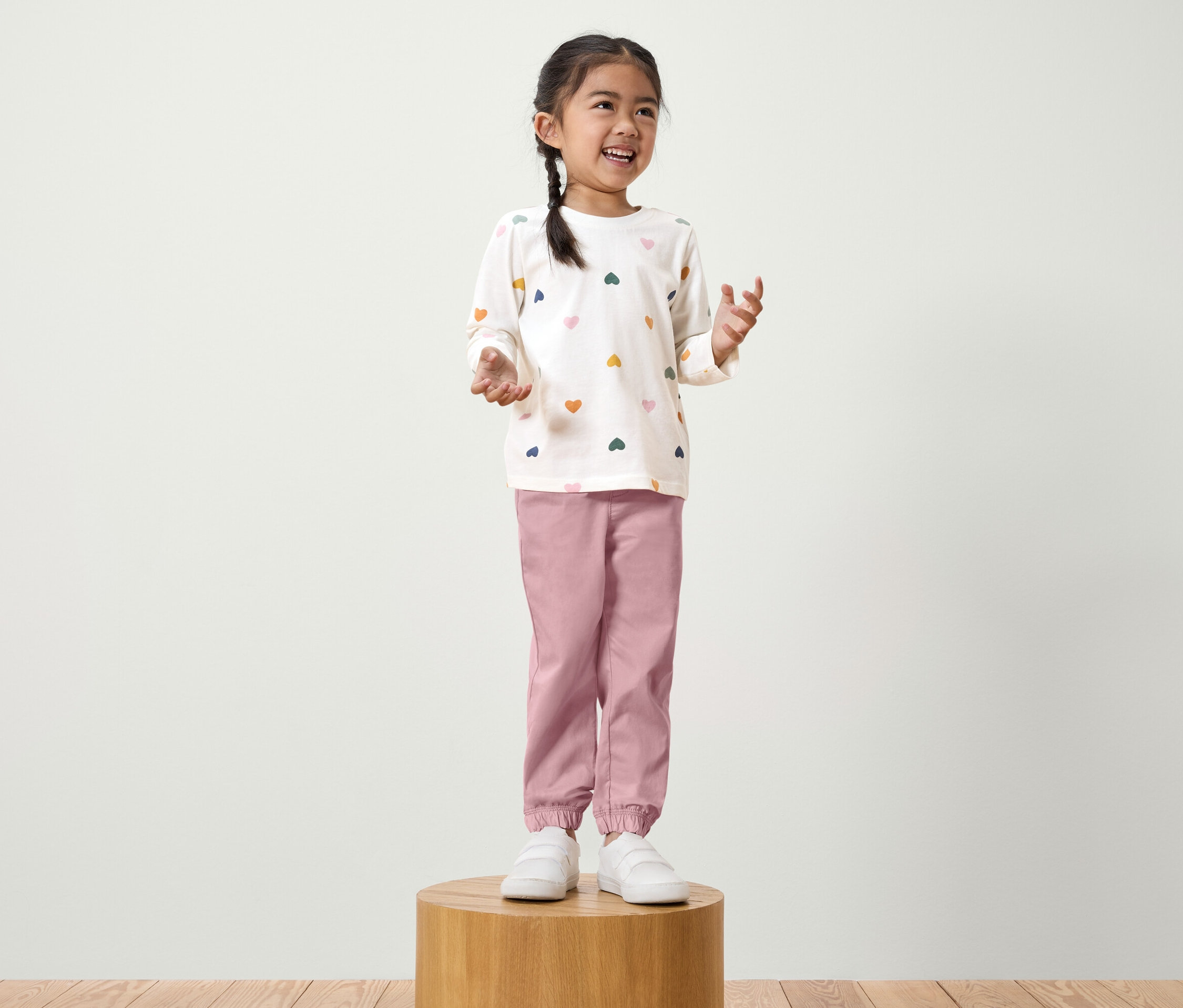 Dievča s vrkočom stojí na drevenom podstavci. Má na sebe biele tričko s dlhým rukávom s farebnými srdiečkami a ružové nohavice.