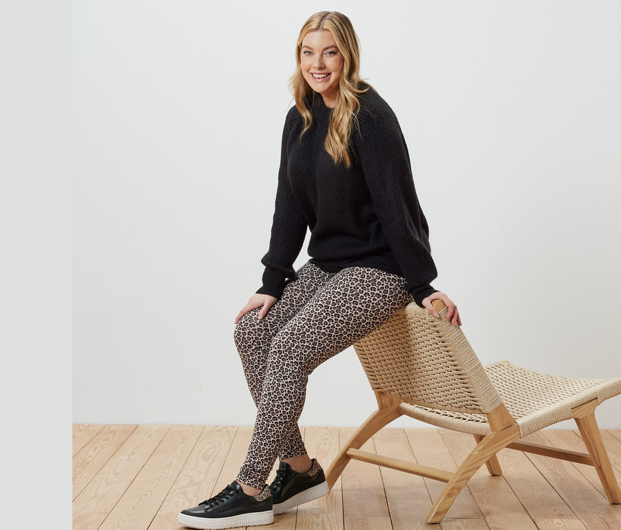 Žena sedí na prútenom kresle a má na sebe čierny pletený pulóver a vzorované legíny.