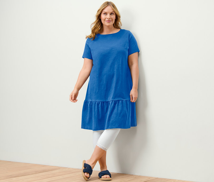 Žena pózuje v kobaltovomodrých džersejových šatách s volánom, bielych legínach a modrých sandáloch.