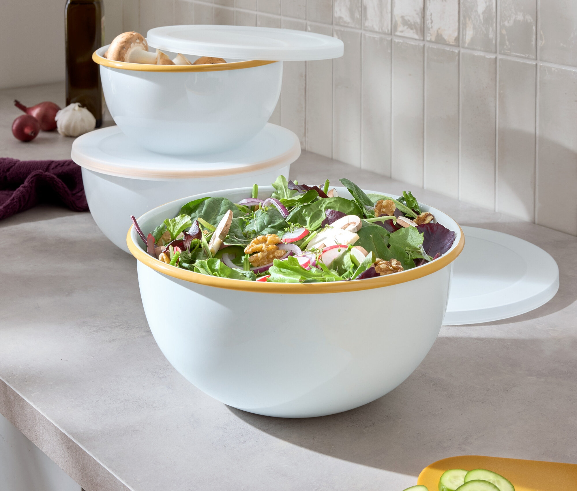 Tri biele misy s farebným okrajom naskladané na seba, najväčšia misa obsahuje šalát, vedľa leží žltá doska na krájanie s nakrájanou uhorkou.