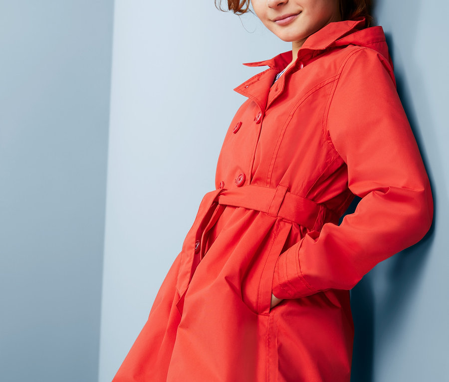 Dievča sa opiera o stenu v červenom trenčkote do každého počasia z recyklovaného materiálu.