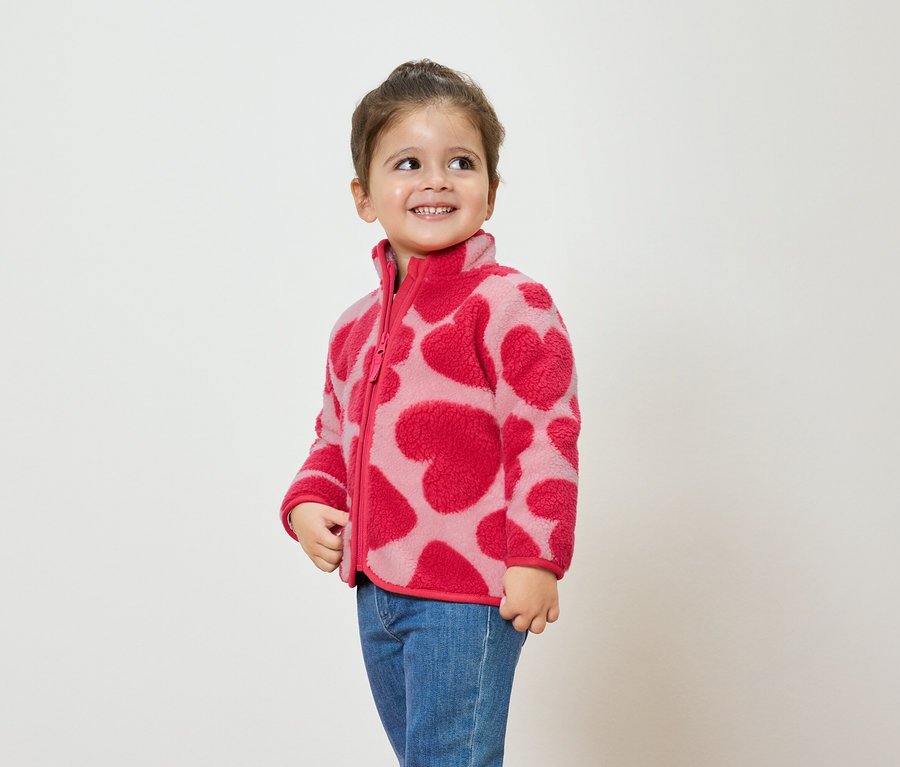 Dievča v ružovej fleecovej bunde so vzorom sŕdc a modrých džínsoch sa usmieva a pozerá sa nabok.