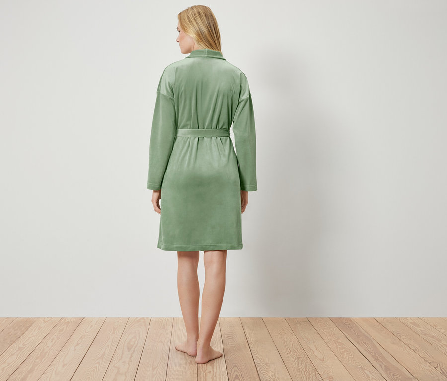 Pohľad zozadu na blondínu v svetlozelenom župane, ktorá stojí na drevenej podlahe pred bielou stenou.