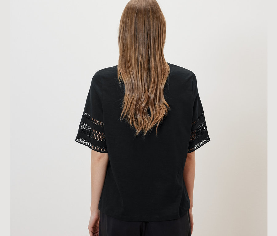 Zozadu je vidieť žena s hnedými vlasmi. Má na sebe čierne tričko s výšivkou a čierne nohavice.